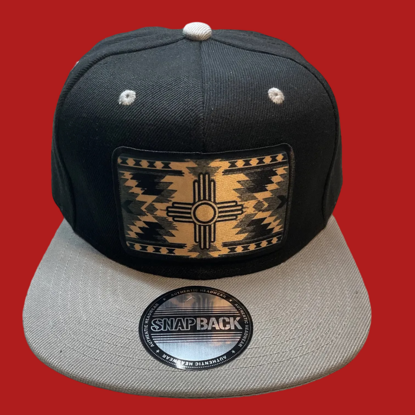 A black and gray snapback cap featuring a rectangular front patch with a Southwestern-style geometric pattern in tan and black. The brim has a circular 'SNAPBACK' sticker, and the hat is photographed on a textured beige fabric surface.