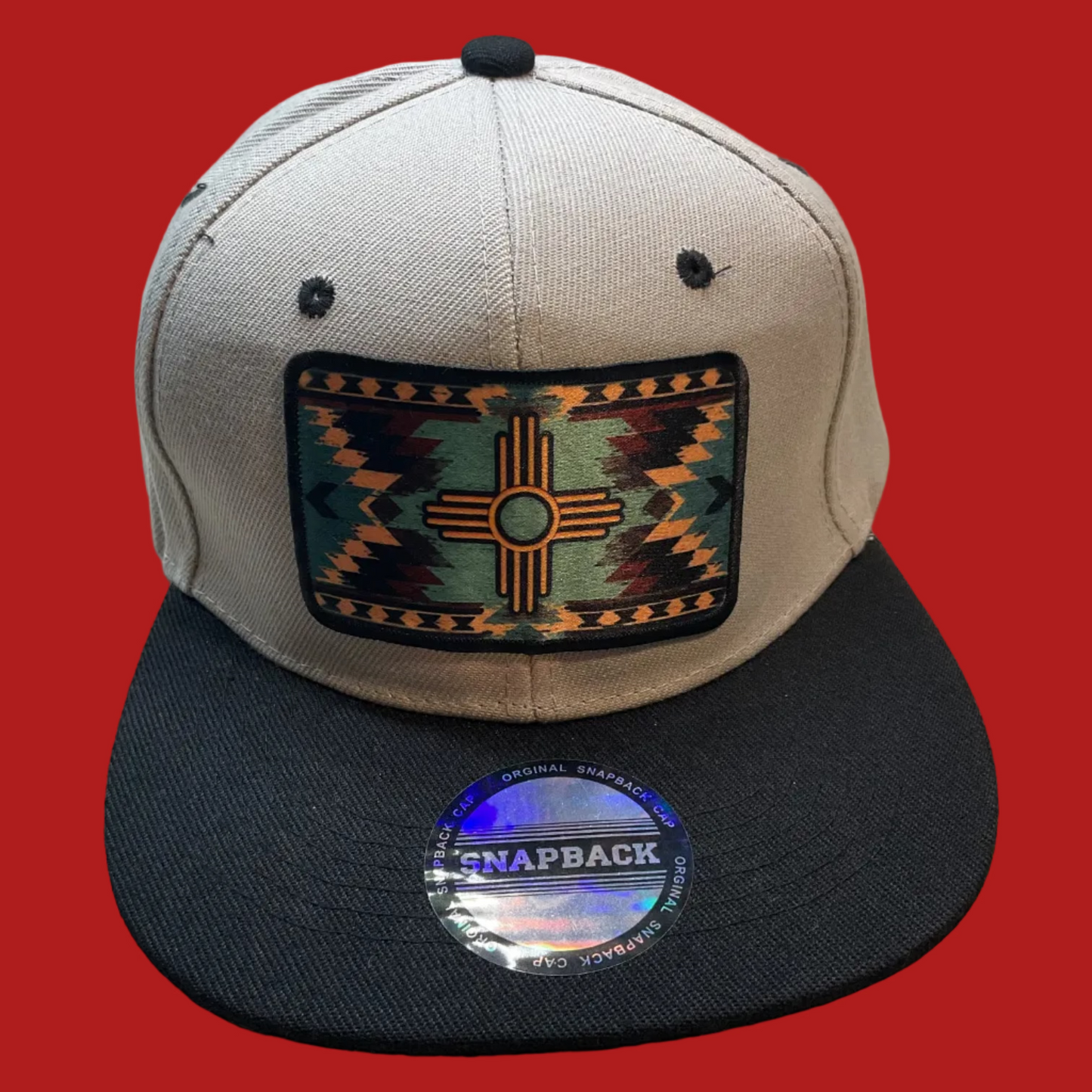 A structured snapback cap with a light gray crown and a black flat brim, featuring a rectangular Southwestern-style geometric patch on the front with a Zuni cross motif. A round holographic 'SNAPBACK' sticker is visible on the brim.